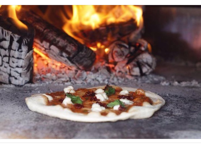 A pizza cooking on the stone floor of a wood fired oven with flames burning behind it