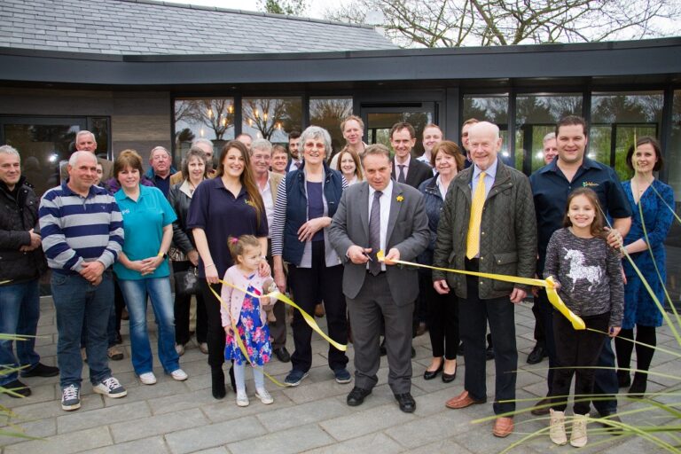 A group gathered outside the new reception building for the ribbon cutting, with a gold ribbon across the doorway