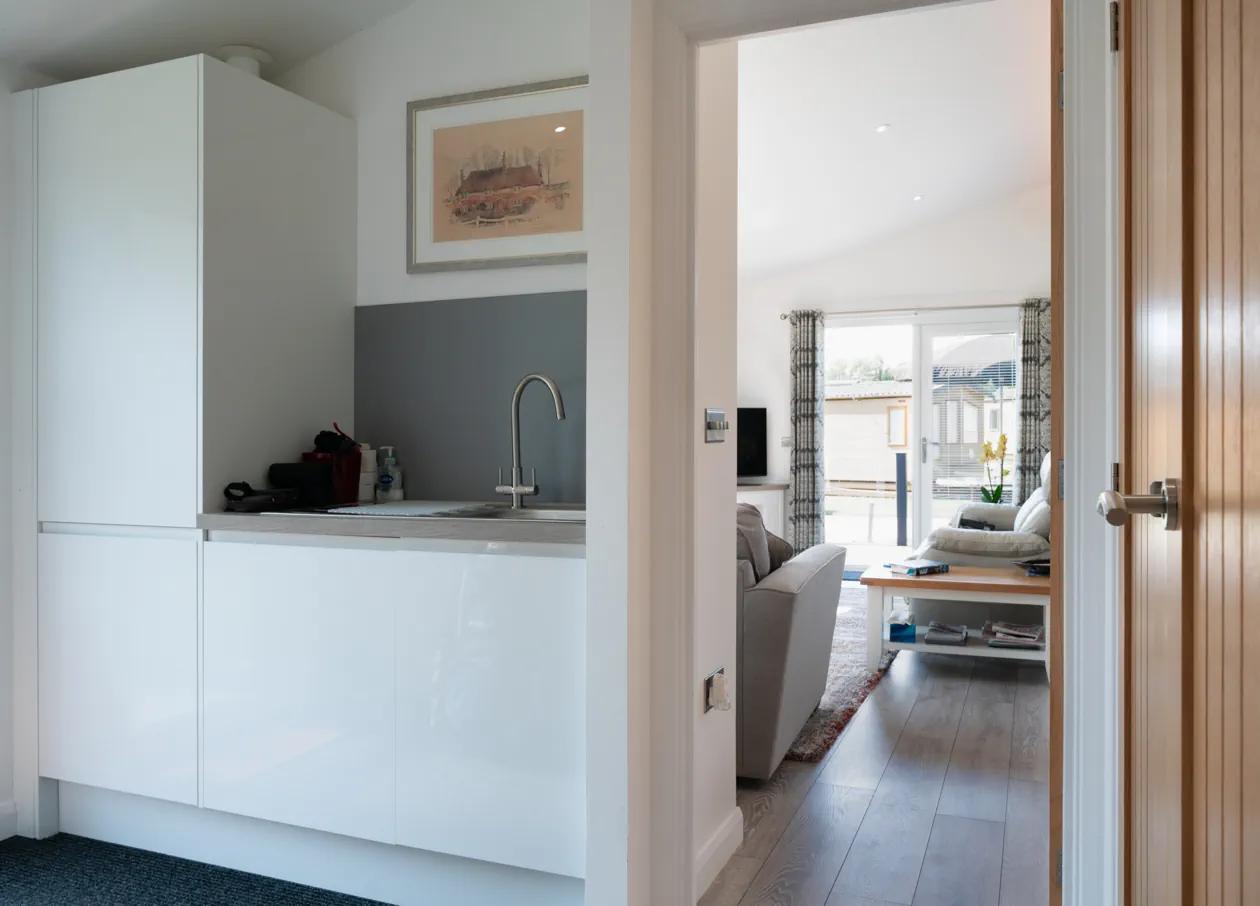 Modern open-plan lounge with stylish sofas, wall-mounted TV, vaulted ceiling, and a view into the sleek fitted kitchen — blending comfort and practicality.