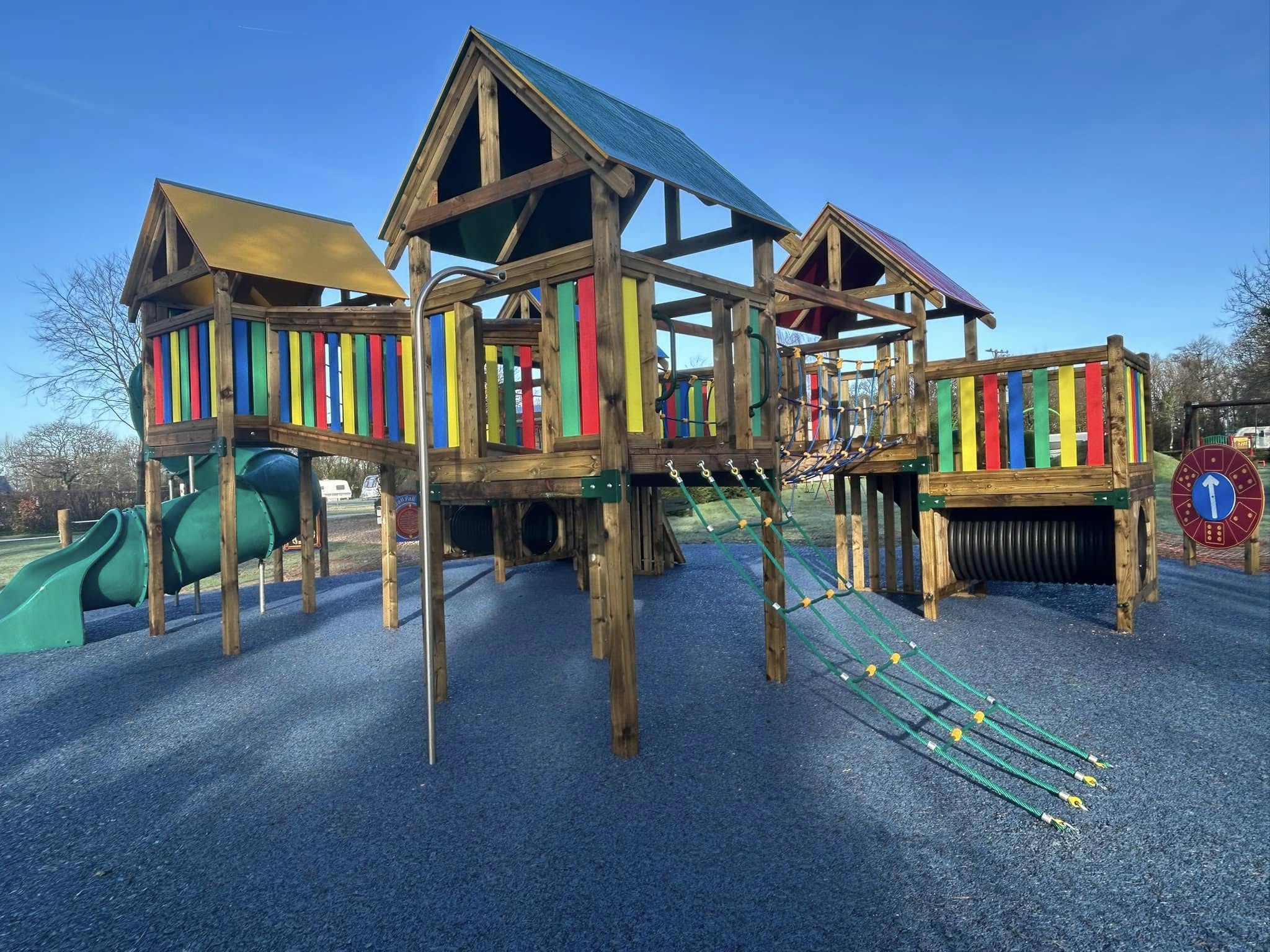 Two wooden climbing frames with slides, ladders and coloured panels on a green safety surface