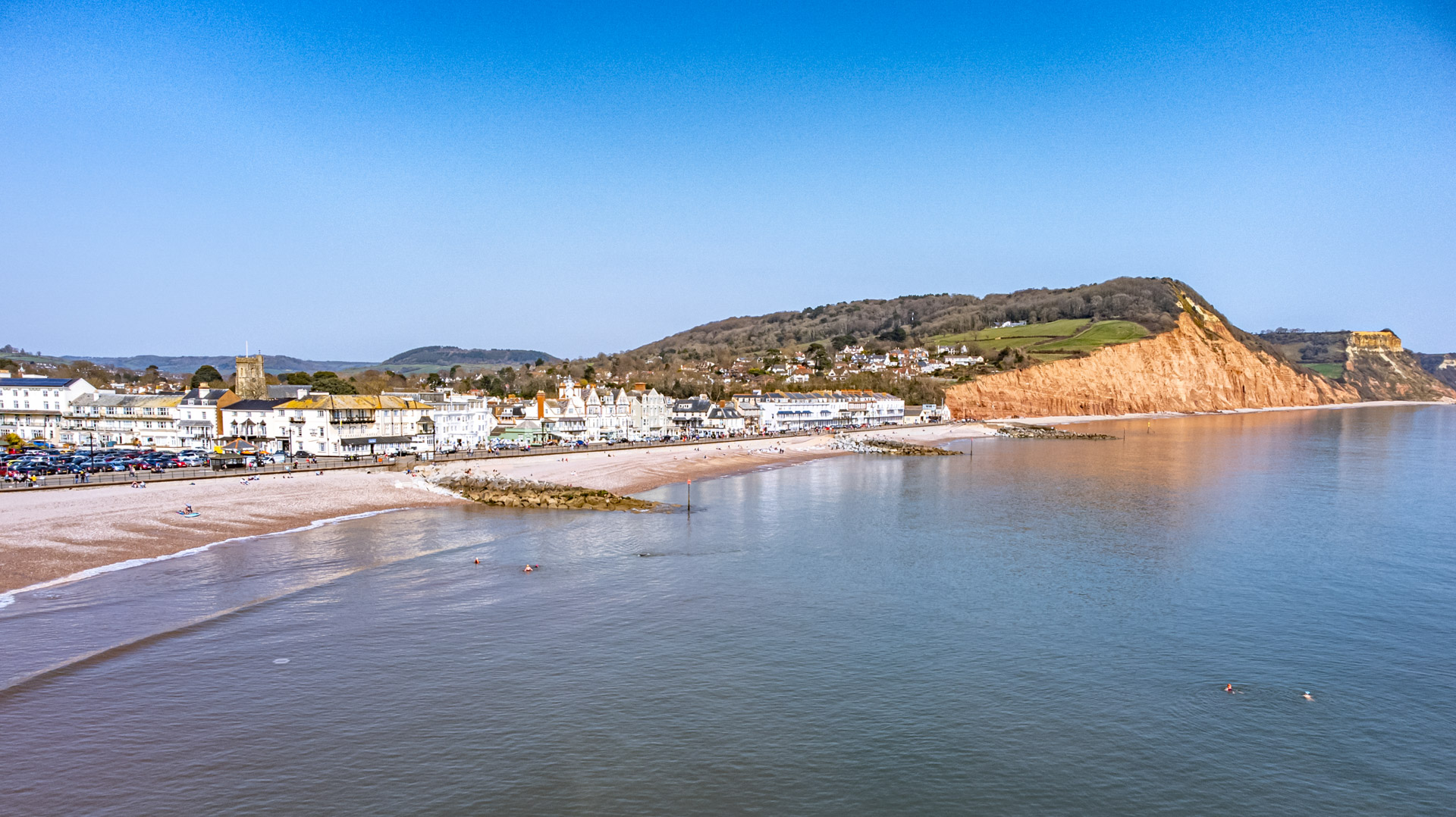 Sidmouth beach from above, the shingle curving away below the red cliffs with the town along the front