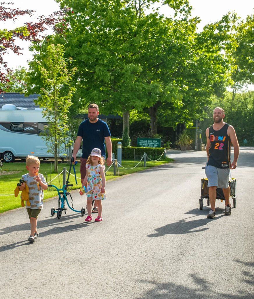 A man pushing a pushchair along the park road with two young children walking ahead of him, hedging and pitches alongside