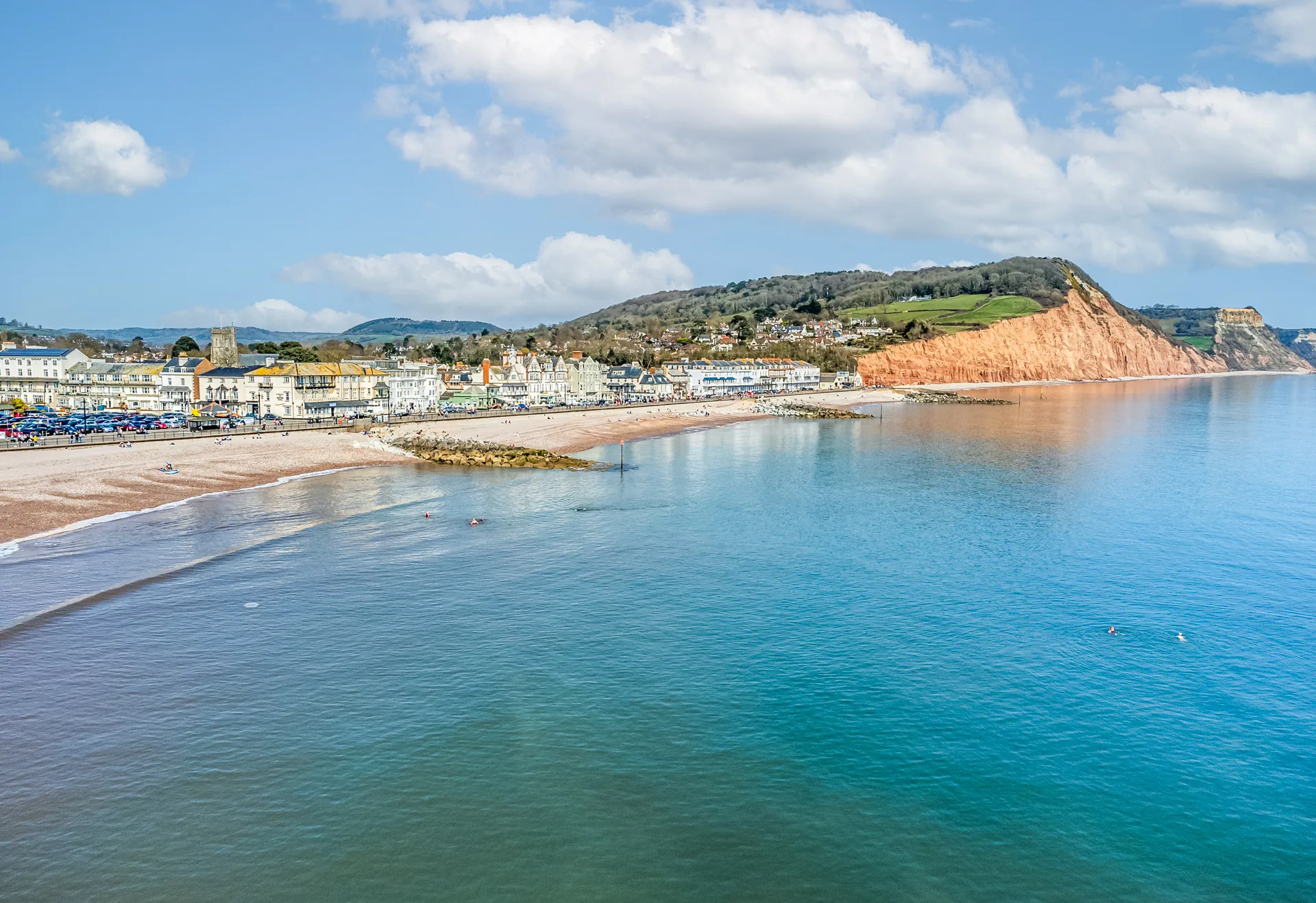 Elevated view along Sidmouth seafront, with the beach, the rock breakwaters offshore and green hills rising behind the town
