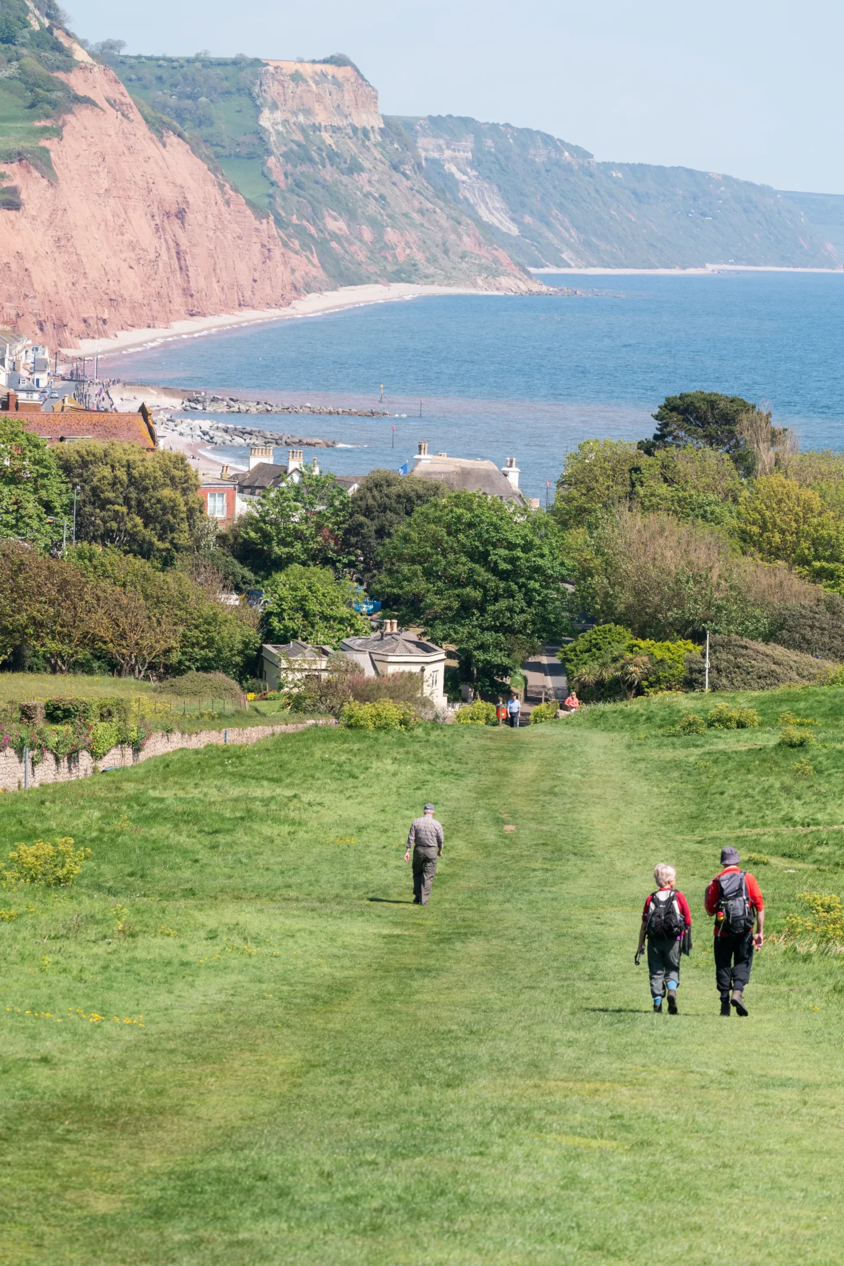 Two walkers on the grassy coast path on Salcombe Hill, with Sidmouth and the red cliffs on the coast behind them