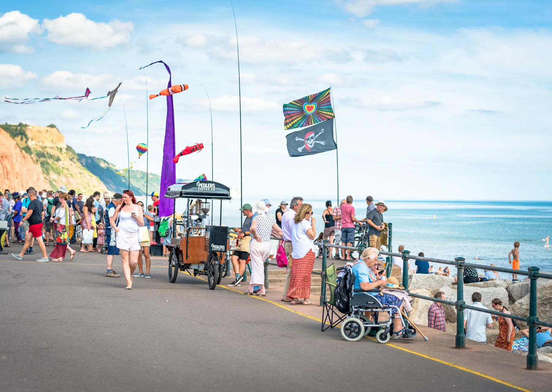 Festival flags and banners flying along Sidmouth seafront, with people walking past a stall and the red cliffs beyond