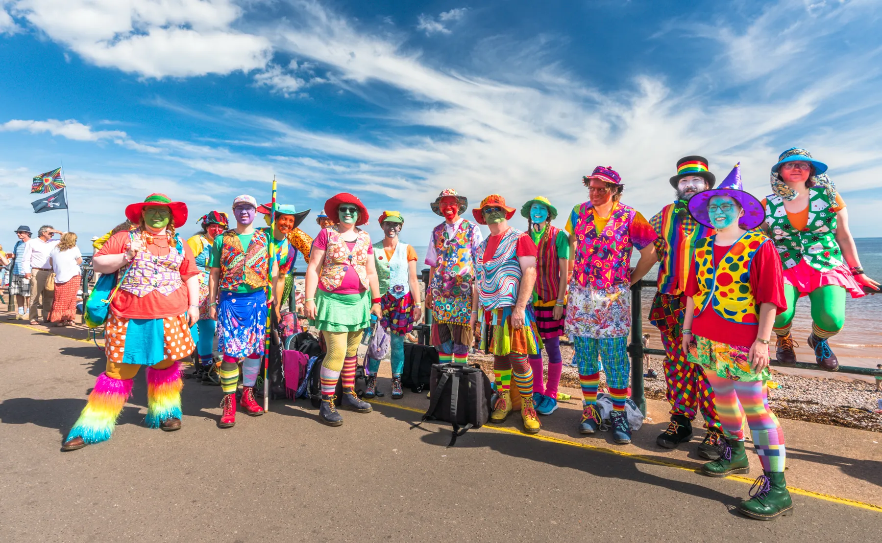 A long line of morris dancers in ragged coats and flowered hats standing along the seafront under a wide blue sky